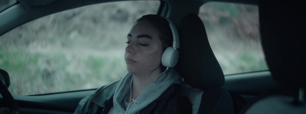 A young woman sits in the drivers seat of a car with her eyes closed wearing large white headphones lost in music