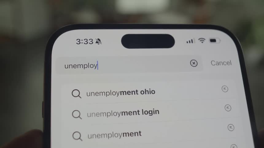 A closeup shot shows a smartphone screen displaying a search bar with the word unemployment and related search suggestions
