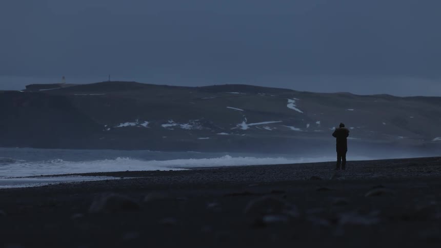 A solitary figure stands on a dark rocky beach as waves crash against the shore under a dusky sky