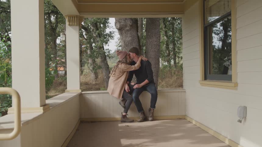 Couple sits on the enclosing half wall of the porch exchanging kisses