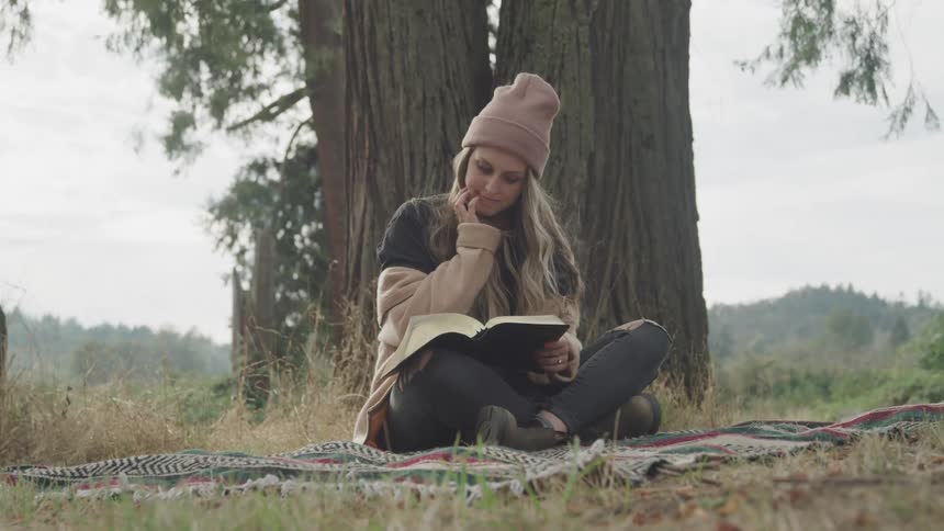 A woman sits by a tree reading a book