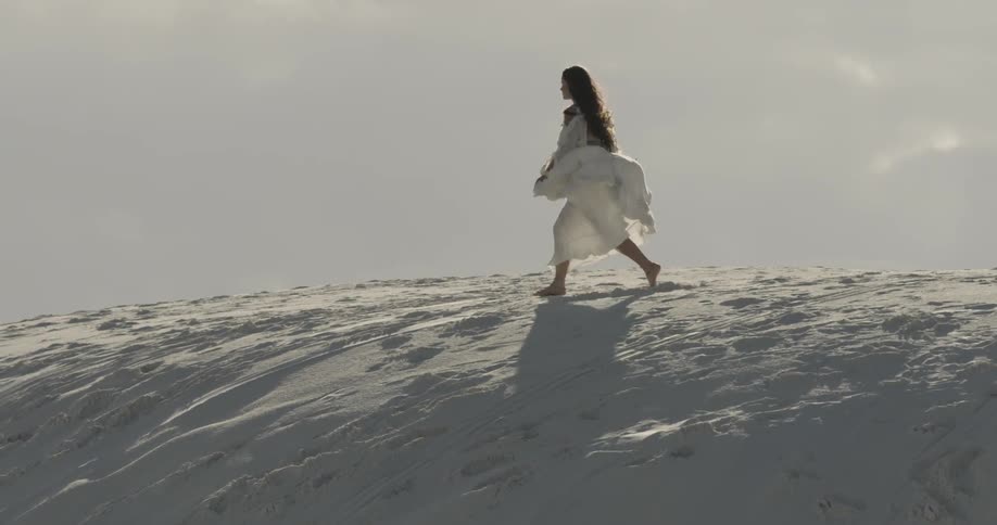 Long-haired woman wearing a long white dress skipping playfully on a hilltop on a cloudy day