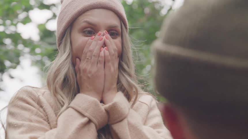 A woman in a beige coat and pink beret clasps her hands over her mouth in surprise as another person appears to kneel before her