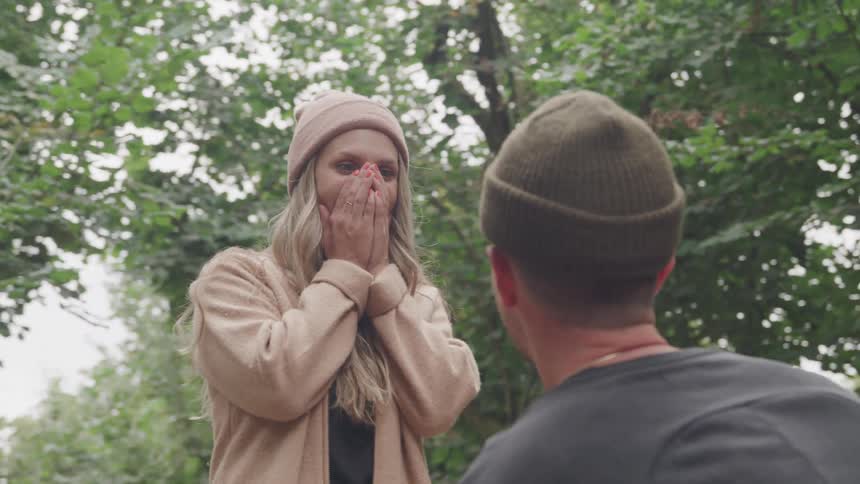 Woman getting surprised and putting her hands on her mouth while a man in front of her is looking on kneeling in a forest
