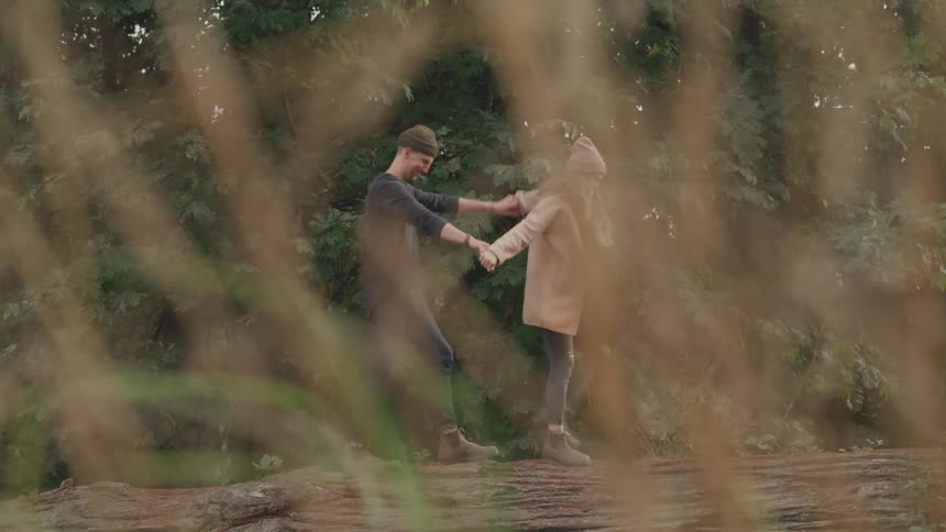 A couple holding hands walk atop a massive fallen tree trunk in a forest surrounded by tall dry grass in the foreground