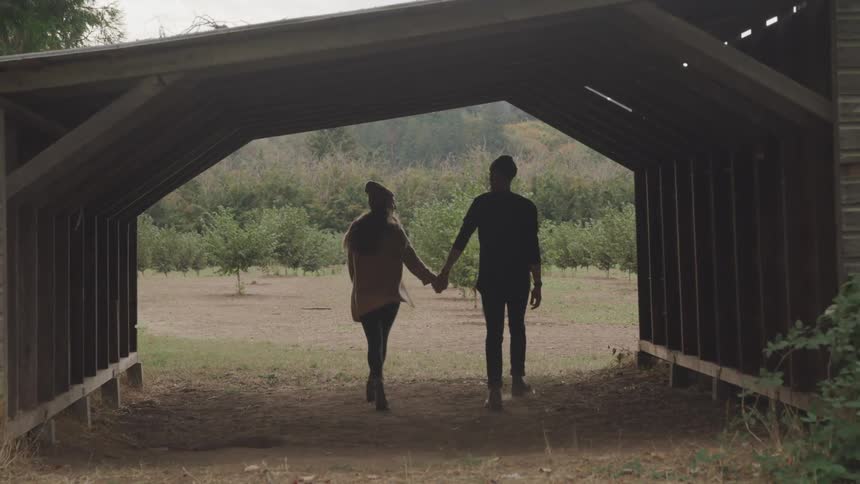 A couple walk hand in hand under a rustic wooden structure heading toward an orchard with neatly aligned rows of small trees