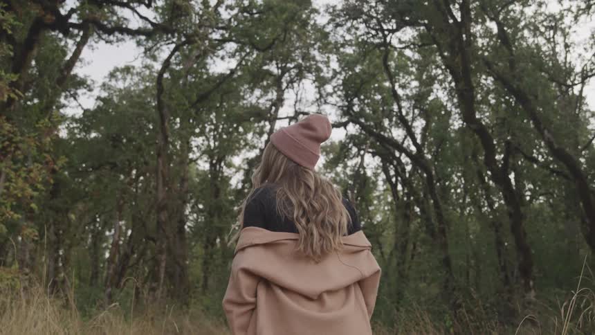 A woman with long hair wearing a black shirt and a beige coat while standing in a forest surrounded by tall trees and dense foliage