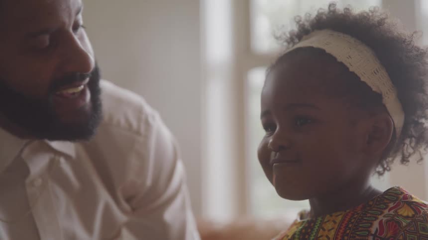 A bearded father and his daughter wearing a white headband and a vibrant patterned outfit share a kiss in a room near a window