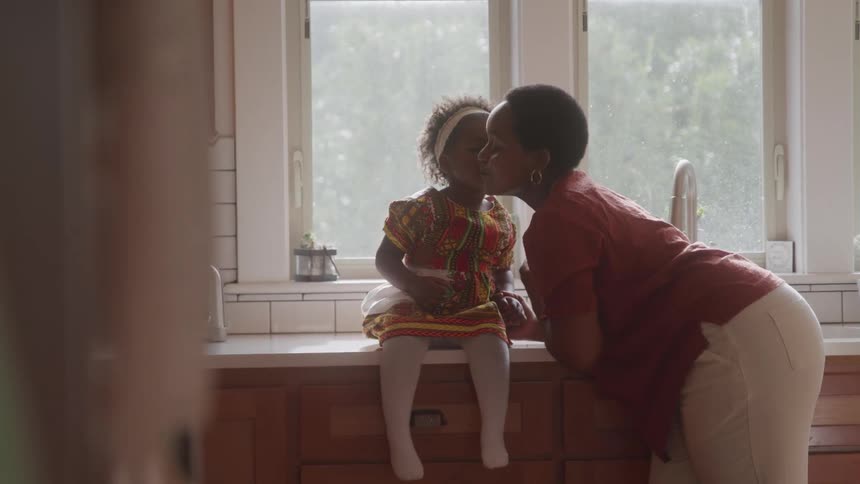 An adult woman next to a little girl sitting next to a sink and getting a kiss on the cheek from her