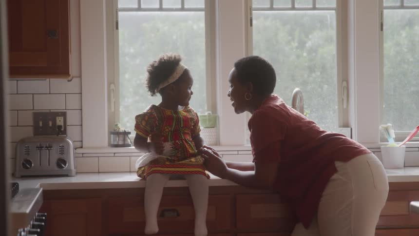 A woman is leaning on a kitchen counter talking to a little girl who is sitting in front of her