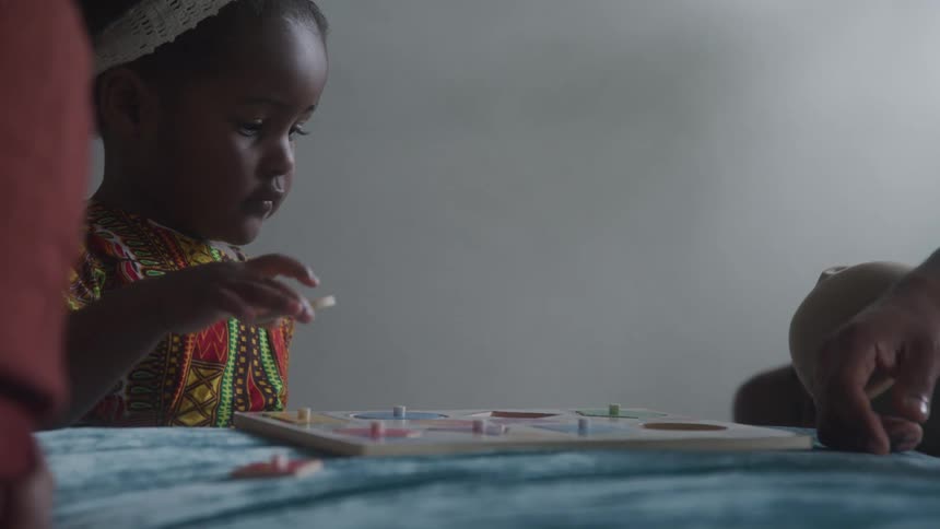Toddler girl solving a wooden puzzle while and adult sits near