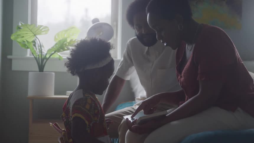 Two parents sit on a bed in a sunlit room teaching their daughter who is dressed in a white headband and a vibrant patterned outfit how to read