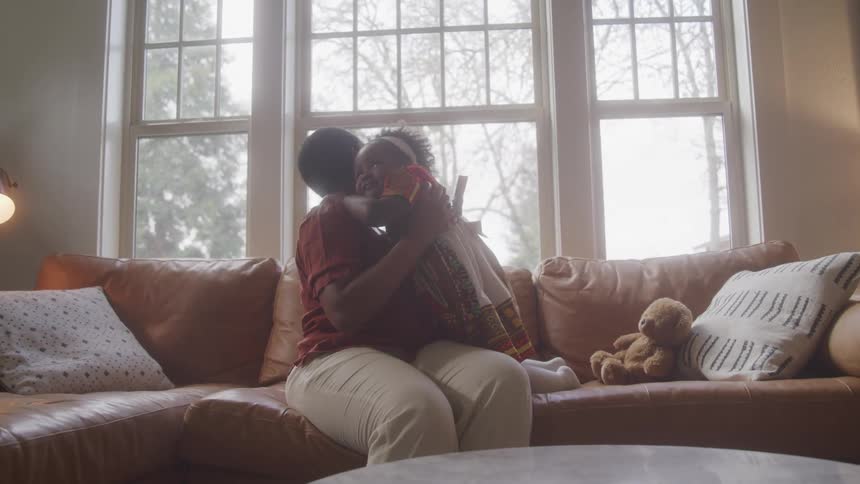 A mother sits on a couch near a window hugging her toddler daughter on a sunny day