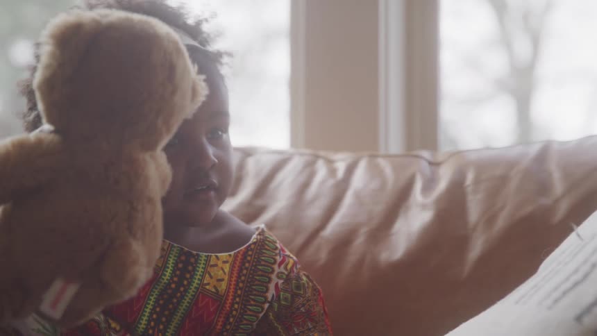 A teddy bear being handed to a little girl and she places it next to her next to a pillow