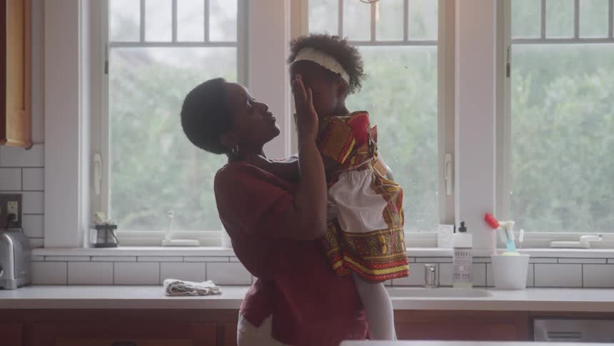 A mother is wiping the tears from the face of her daughter who is on her lap crying in a kitchen