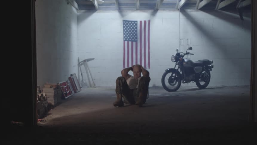 A man in militarystyle clothing is doing situps in a dimly lit garage with an american flag hanging on the wall behind him and a motorcycle parked nearby creating a gritty and determined atmosphere