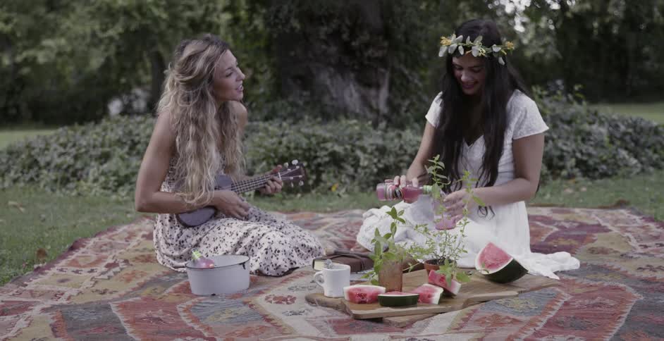 One woman pours a drink and another plays a ukulele during a picnic