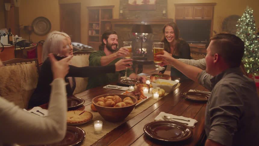 A group of people gathers around a wooden dining table raising glasses in a toast with a christmas tree and a fireplace in the background