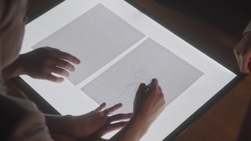 Three individuals work at a light table with one person drawing on a sheet of paper using a pen