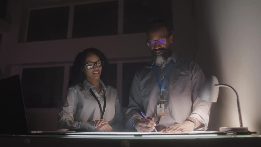 Two individuals stand at a desk with a laptop and a desk lamp reviewing documents under soft dim lighting