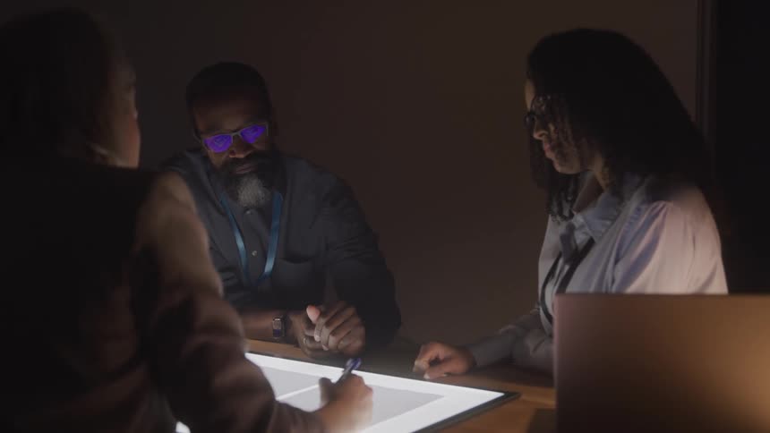 Three individuals stand around a desk reviewing documents illuminated by soft dim light coming from the desk