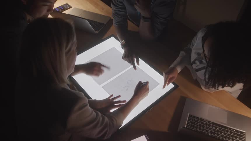 A group of people work at a light table with one person drawing on a sheet of paper using a pen