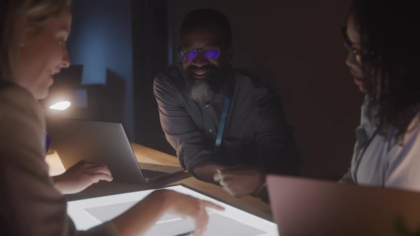 A group of people stands around a light table in a dimly lit room discussing while laptops and papers are spread across the table