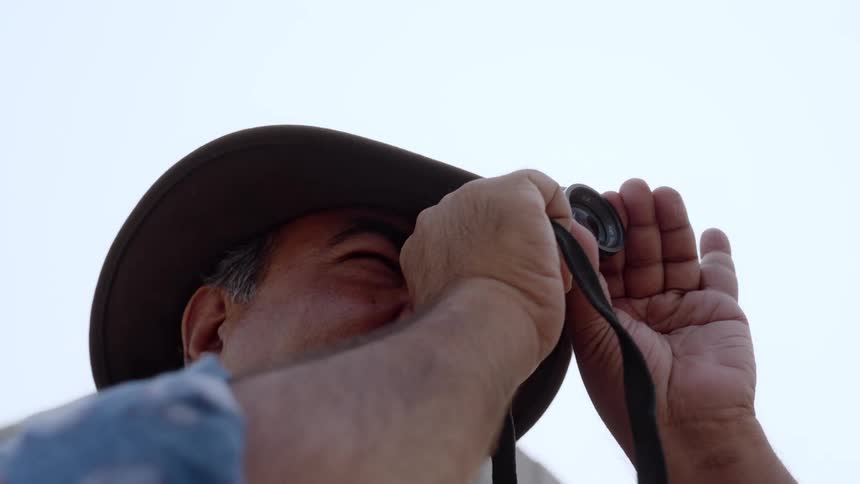 A man wearing a hat and holding a camera looks upwards taking a photograph of something in the distance