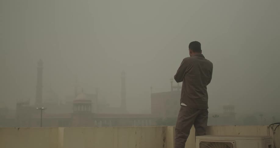 A man stands on a rooftop overlooking a hazy cityscape likely photographing the scene