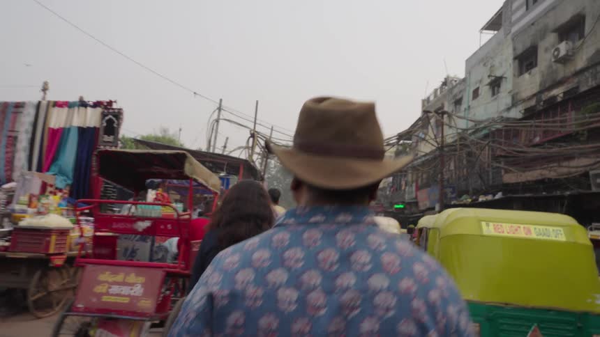 A person in jeans and brown shoes walks away from a green auto rickshaw on a dusty city street