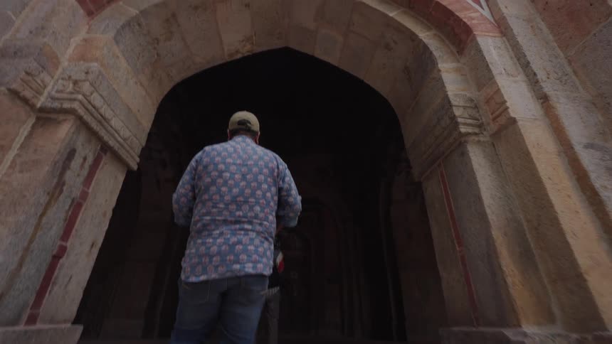 A person wearing a hat and patterned shirt walks forward into an ornate stone archway
