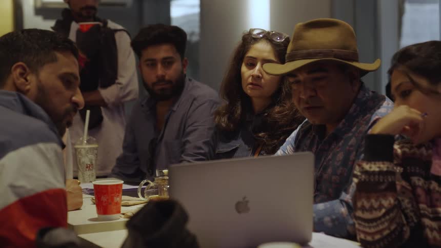 A group of people sits around a table with a laptop drinks and papers actively engaging in a discussion