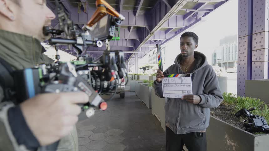 A film crew prepares for a shoot beneath a bridge as a person clicks a clapboard in front of the camera