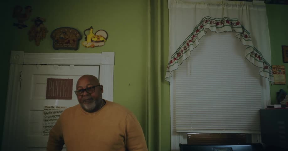A man wearing a yellow sweater standing on a room at home talking to someone