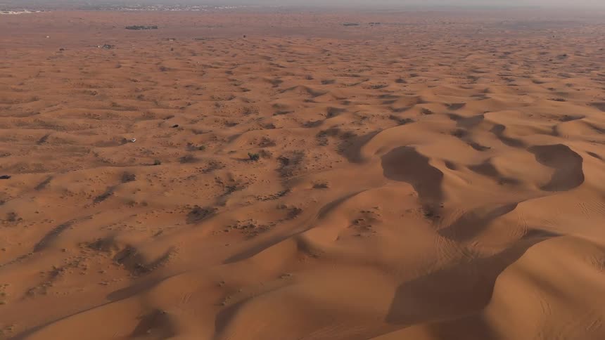 A vast undulating desert landscape stretches to the horizon under a hazy sky marked by subtle tire tracks and two small vehicles one black and one white driving across the terrain