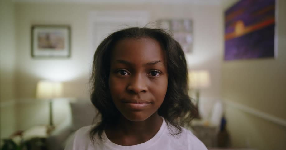 A young girl with a gentle smile stands in a warmly lit living room looking directly into the camera with a calm and confident expression