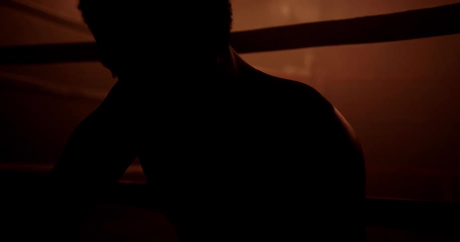 The silhouette of a shirtless man sits beside a boxing ring in a dimly lit gym