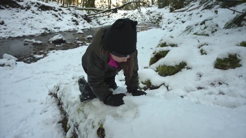 Little girl climbing snow coverd fallen tree in forest near a creek