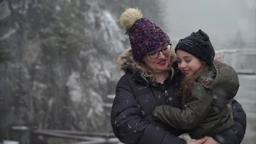 A mother holding her daughter in her arms both dressed in winter clothing stand on the snowy deck of a ship gazing overboard