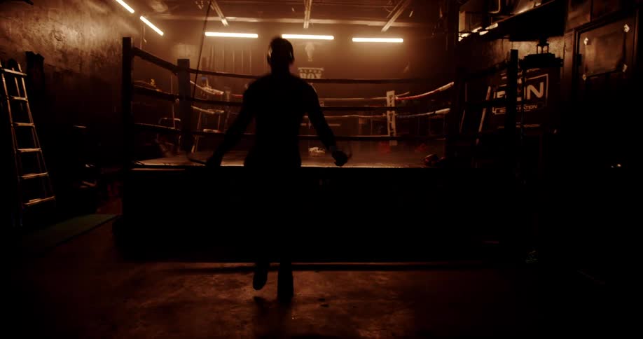 A silhouette of a boxer jumps rope near a ring in a dimly lit gym