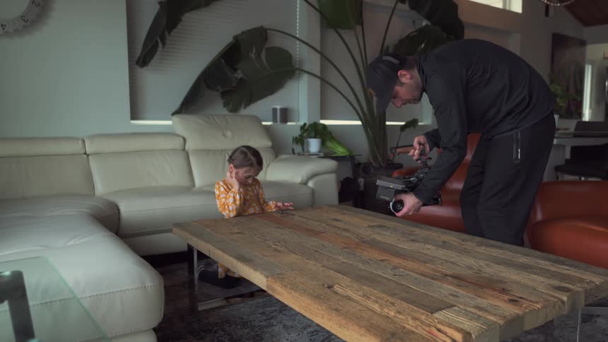 The man holding the camera is filming the little girl who is sitting at the table playing with a toy