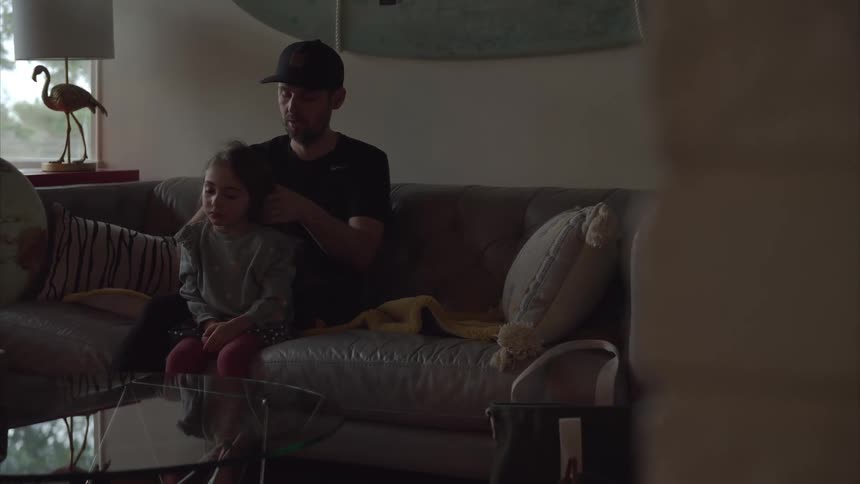 A man and his daughter are sitting on a couch as he brushes her hair