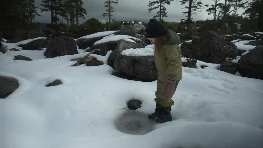 A girl wearing winter clothes plays with a frozen puddle of ice surrounded by snow