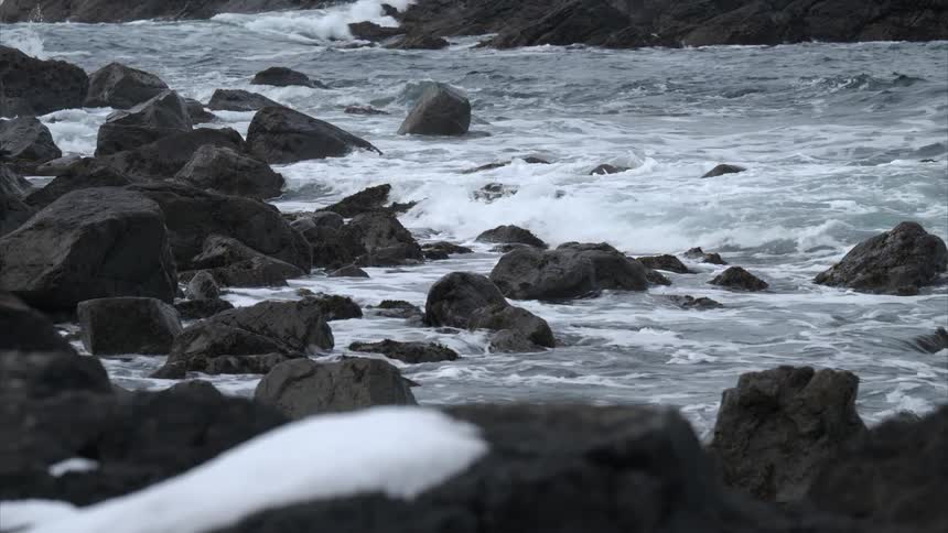 Whitecapped waves roll onto a rugged shoreline strewn with dark rocks and light snow patches as foamy water swirls between the stones and the subdued coastal setting stretches across the frame