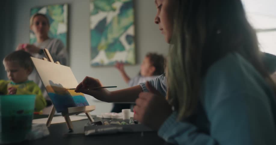 A girl paints on a small canvas set on an easel surrounded by art supplies and a green container on a table