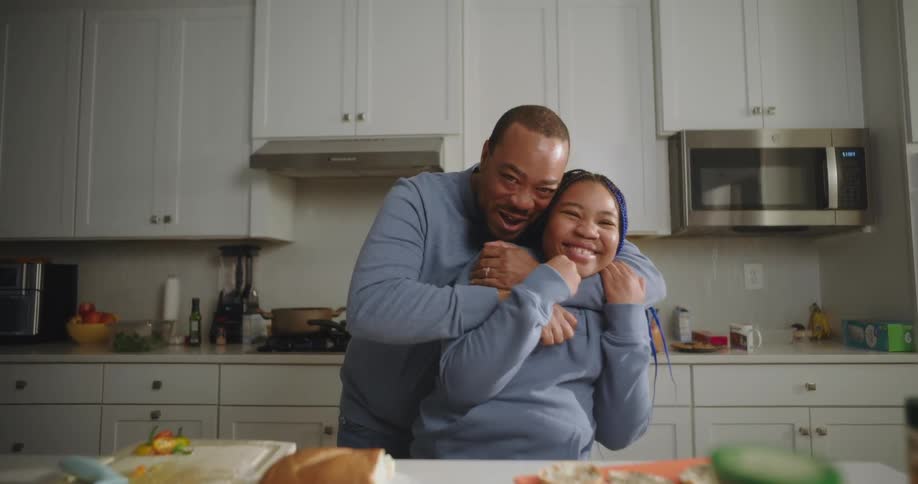 A man embraces and playfully kisses a young woman in a kitchen both smiling