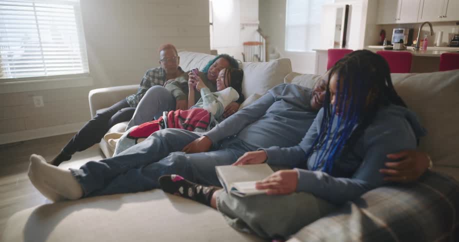 A family relaxes on a large couch in the living room reading and browsing on their phones