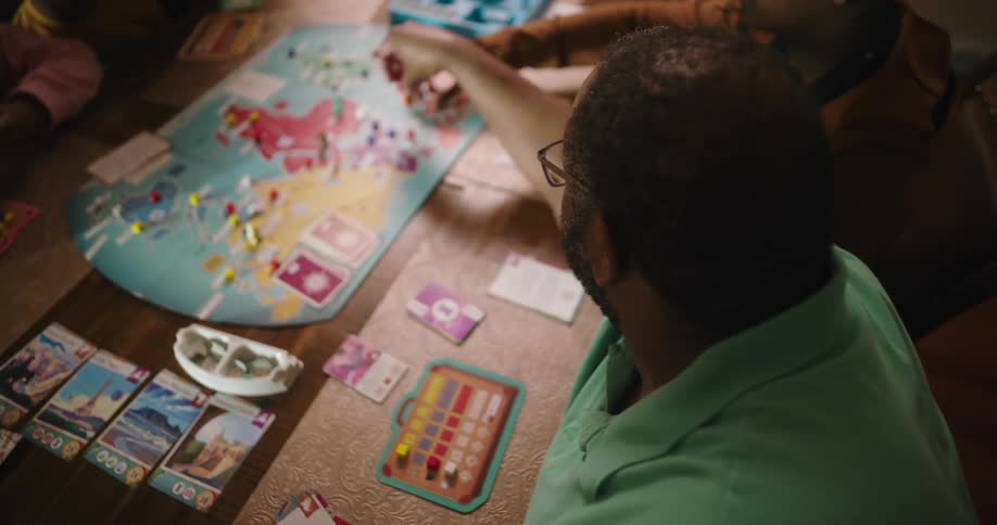 The man and the person beside of him are moving pieces on the game board that is on the table in front of them