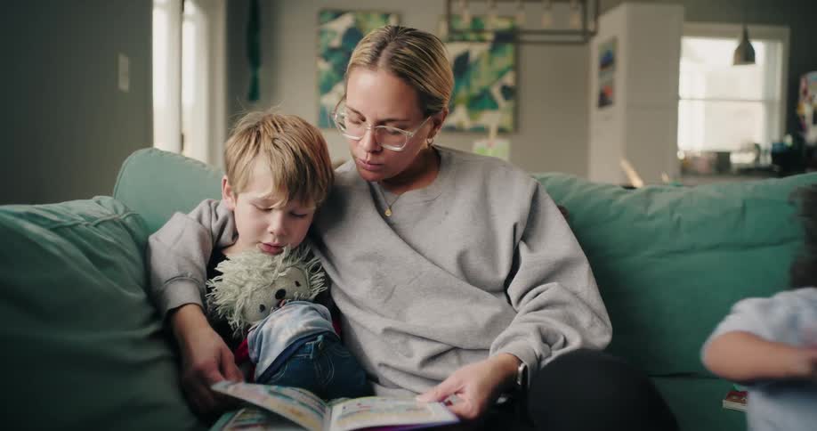 Mother sitting with her young son on the living room sofa reading a book