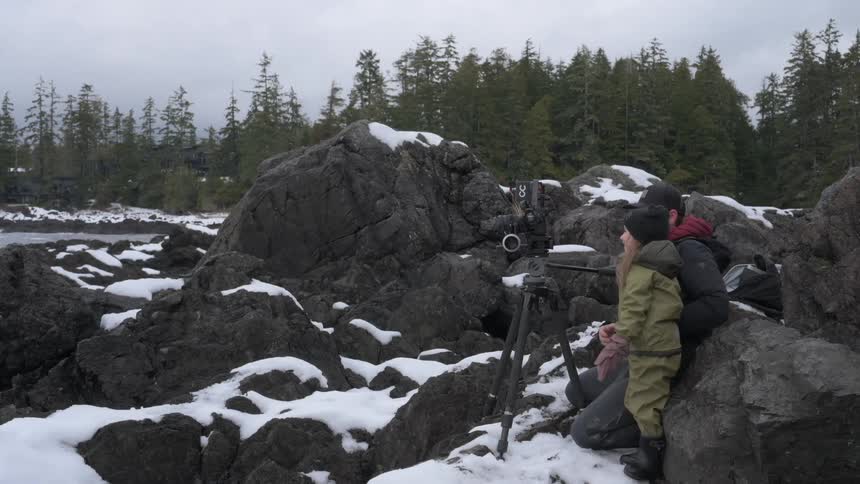 Little girl using a professional camera next to her father while standing in a rocky environment with snow and a forest in the background